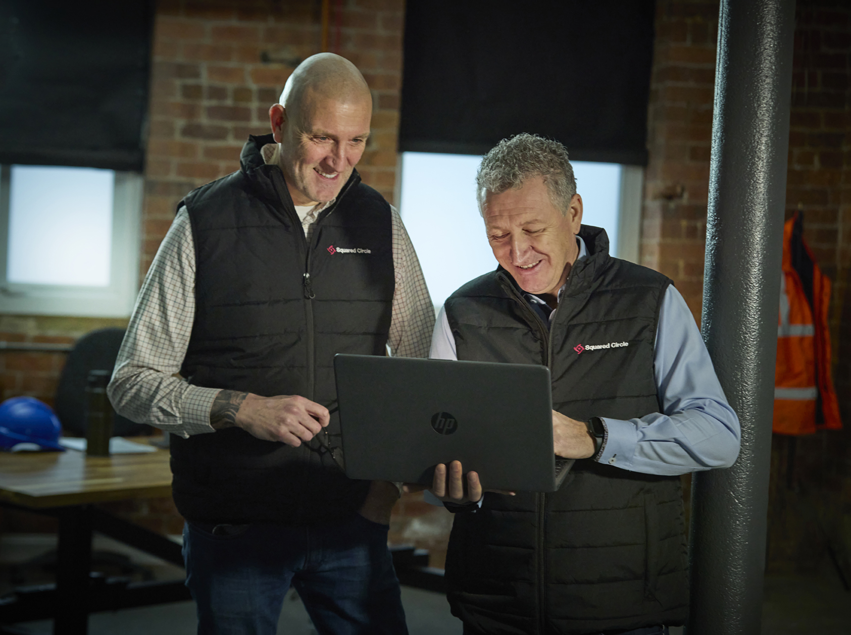 2 males looking at a laptop in an office with an orange vest and a pillar on the background