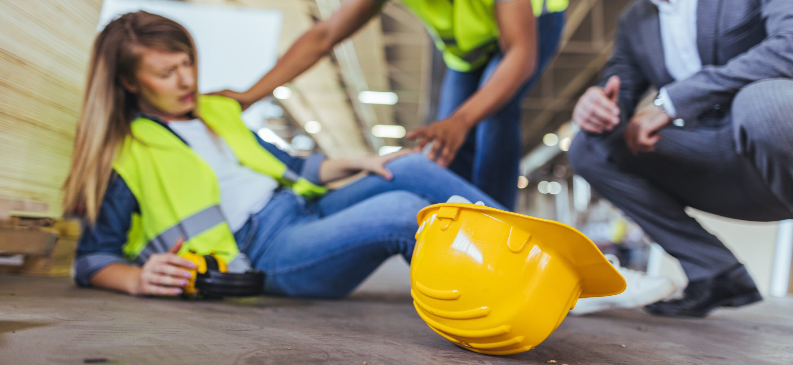 A female worker in a high-visibility vest sitting on a warehouse floor holding her knee after an accident, with a yellow hard hat in the foreground and two colleagues crouching down to assist her.